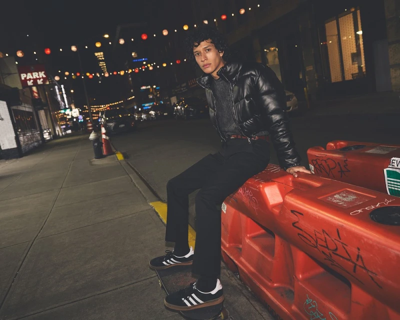 A person sitting on an orange barrier at night on a city street wearing black pants and shoes, a black puffer jacket, and a grey sweater.