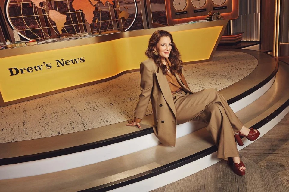 Drew Barrymore sits on a news desk in a beige suit with red platform heels, smiling in a studio setting. News theme.
