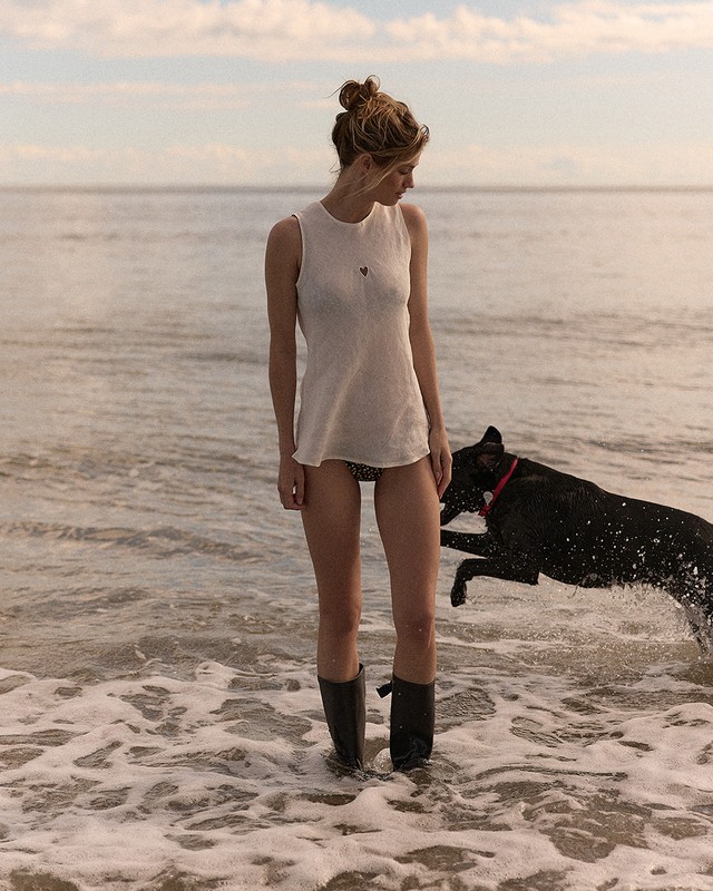 Young woman in a white tank top and black boots stands in shallow ocean water with a black dog jumping nearby against a horizon background.