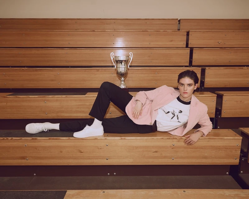 Woman in blazer and athletic shoes lying on bleachers with trophy. Indoor fashion editorial featuring a vintage-style sport setting.