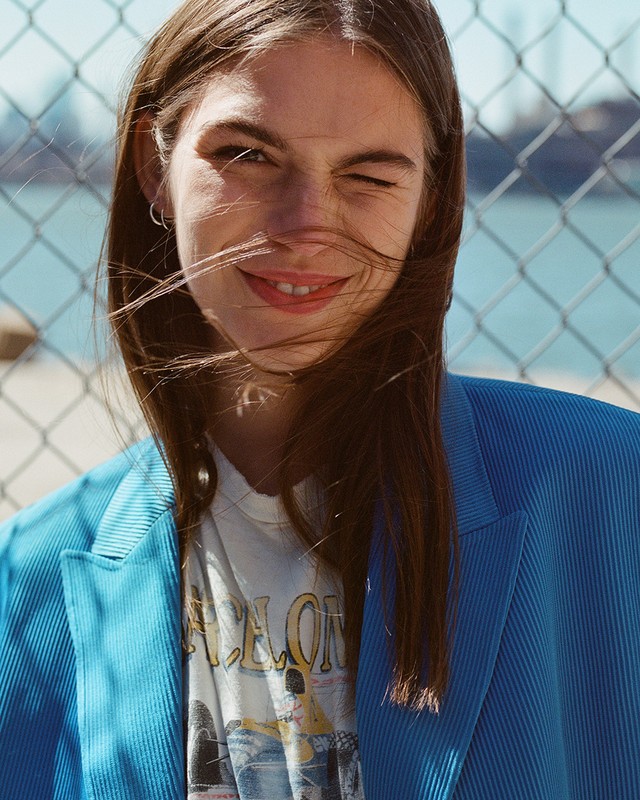 Portrait of a smiling woman with winking expression wearing a blue blazer and graphic t shirt outdoors against a chain link fence