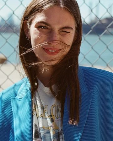 Portrait of a smiling woman with winking expression wearing a blue blazer and graphic t shirt outdoors against a chain link fence