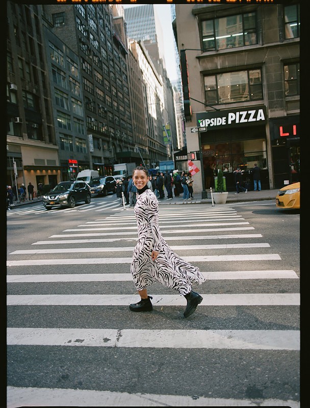 A woman in a zebra-print dress crosses a New York City street on a sunny day, framed by buildings and vehicles.
