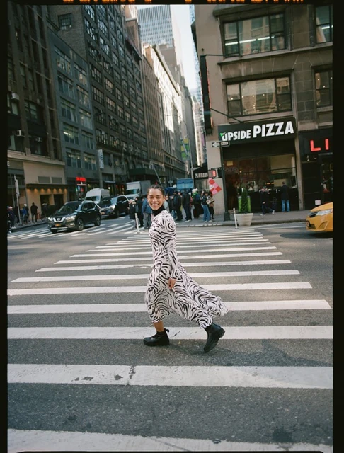 A woman in a zebra-print dress crosses a New York City street on a sunny day, framed by buildings and vehicles.