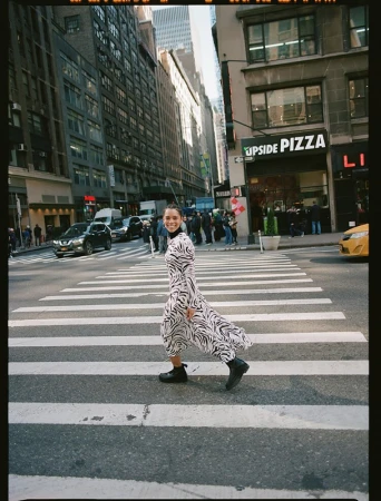 A woman in a zebra-print dress crosses a New York City street on a sunny day, framed by buildings and vehicles.