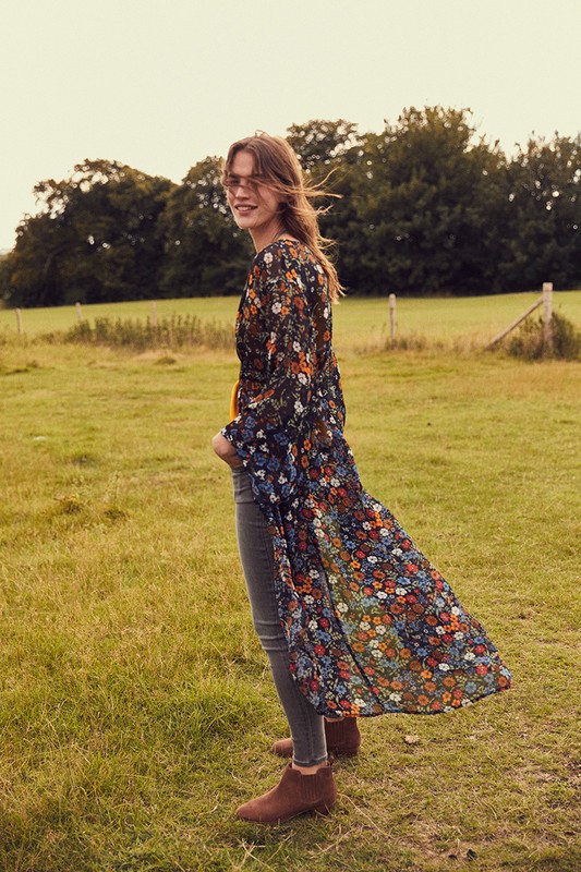A woman in a floral duster, grey jeans, and brown booties stands in a sunny, grassy field with trees in the background.