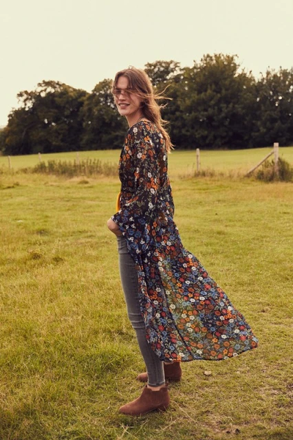 A woman in a floral duster, grey jeans, and brown booties stands in a sunny, grassy field with trees in the background.