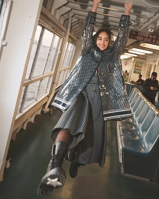 Woman in a shimmering coat playfully hangs from the overhead bar of a ferry, with water visible through the window in the background.