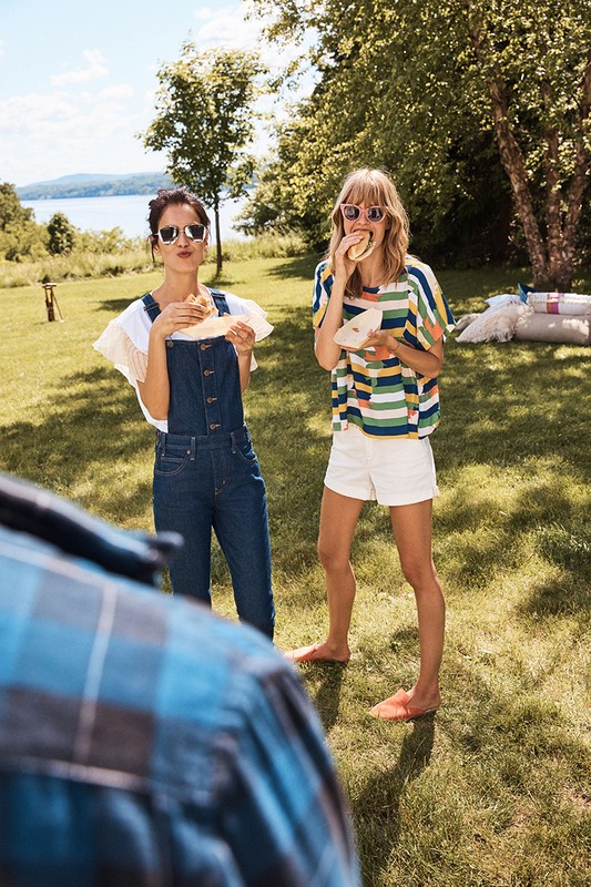 Two women enjoy sandwiches on a sunny day. One in overalls and sunglasses, one in a striped shirt, short-shorts, and blonde hair.
