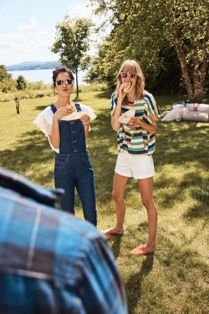 Two women enjoy sandwiches on a sunny day. One in overalls and sunglasses, one in a striped shirt, short-shorts, and blonde hair.