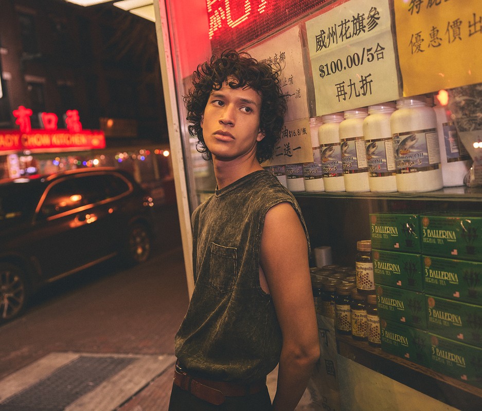 Young adult stands in a dimly lit urban scene near shelves with various products, with neon signs and a passing vehicle in the background.