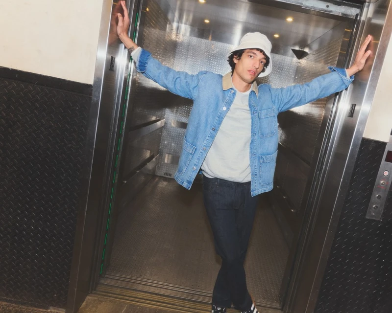 A young adult man wearing a denim jacket and bucket hat stands in an elevator with his arms raised in an indoor environment.