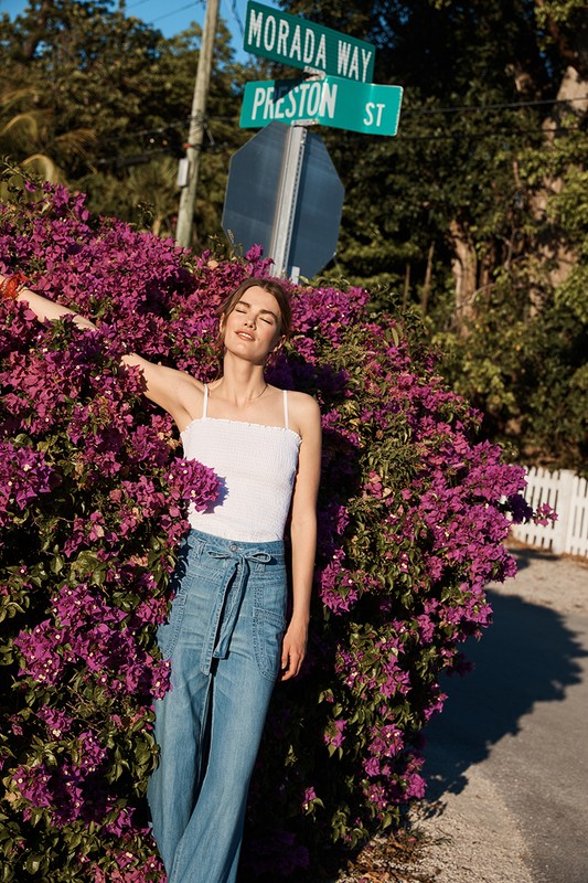 A woman in casual clothing leans against a purple bougainvillea bush near a street sign on a sunny day, showcasing fashion and nature.
