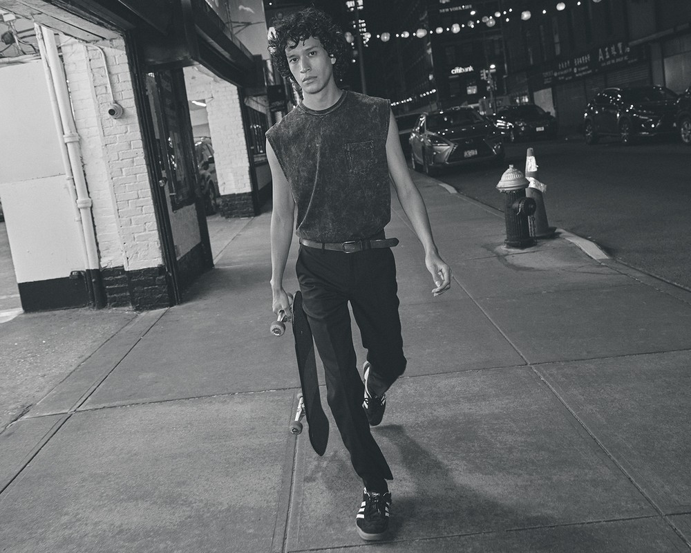 Monochrome medium shot of a young adult male walking with a skateboard at night on an urban street with storefronts and passing cars.