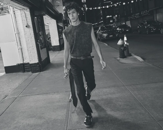 Monochrome medium shot of a young adult male walking with a skateboard at night on an urban street with storefronts and passing cars.