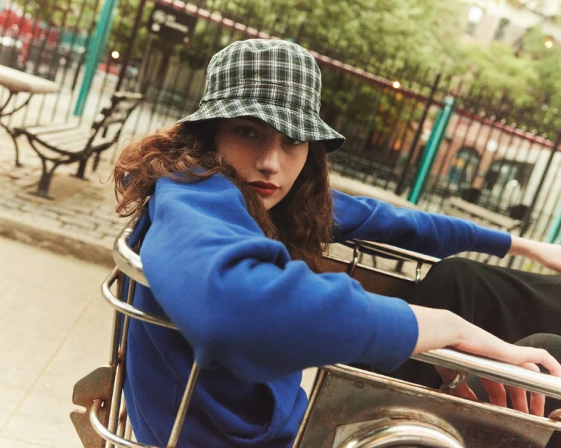 Young woman in a blue sweatshirt and a plaid bucket hat sits in a metal cart in an urban outdoor setting with building background.