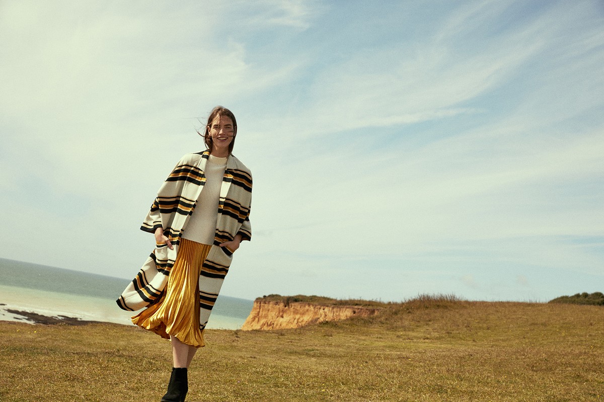 Smiling woman in a striped coat and yellow pleated skirt walking across a grassy coastal cliff under a blue sky.