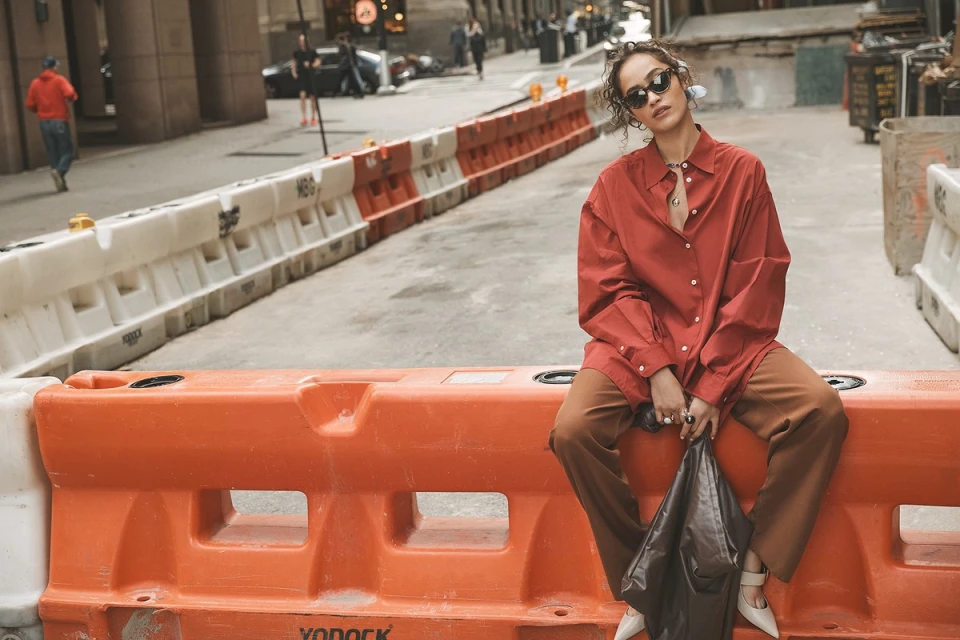 Adult female in red shirt and brown pants sitting on orange construction barrier in urban street with buildings and cars in background.