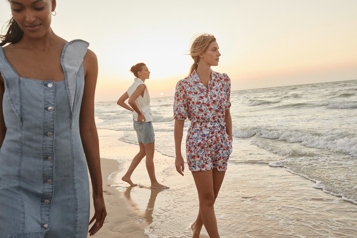 Three women walk along a beach at sunset, wearing summer dresses and separates, with a calm sea and horizon in the background.