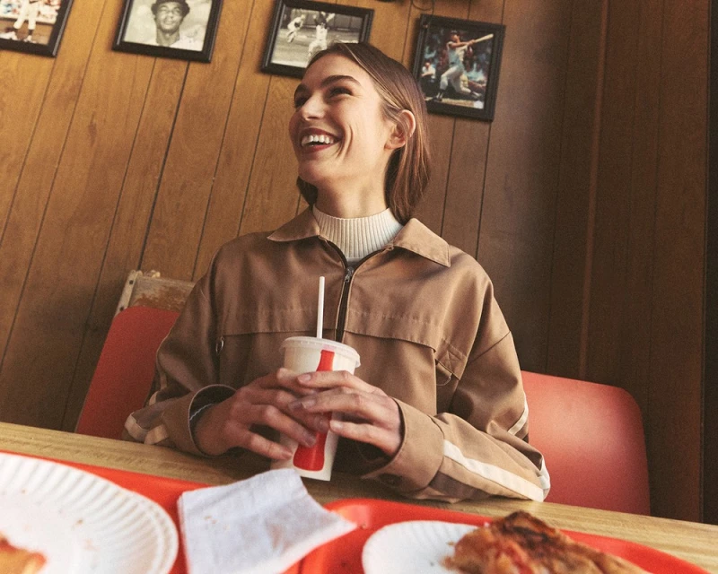 Smiling woman in a brown jacket sitting at a restaurant table with a drink and dishes, with framed photos on the wood-paneled wall.