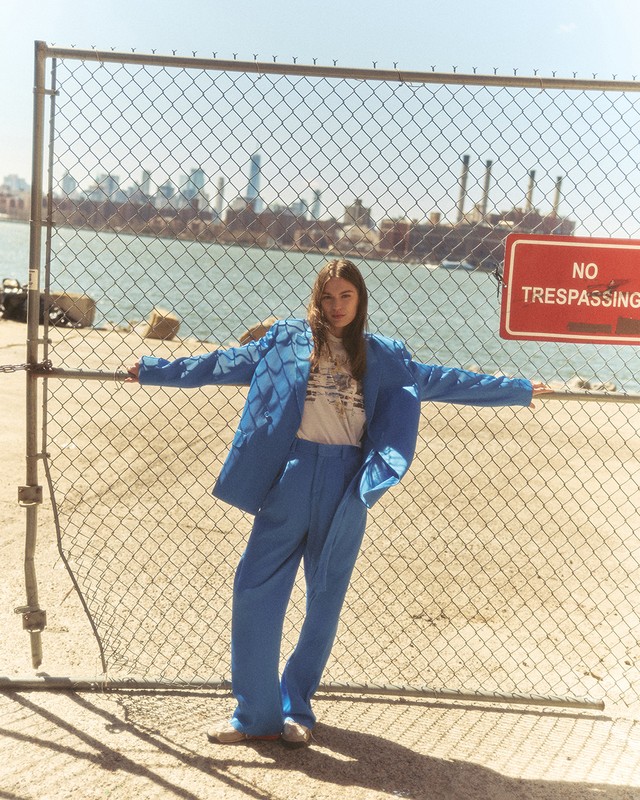 A young adult in a blue suit poses against a chain-link fence with a cityscape and waterway background under bright sunlight.