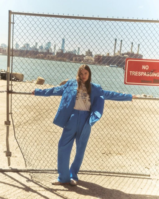 A young adult in a blue suit poses against a chain-link fence with a cityscape and waterway background under bright sunlight.