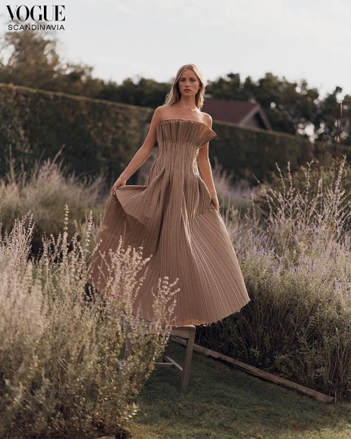 Woman in elegant strapless beige gown stands on wooden stool outdoors amid greenery, Vogue Scandinavia logo in upper left corner.