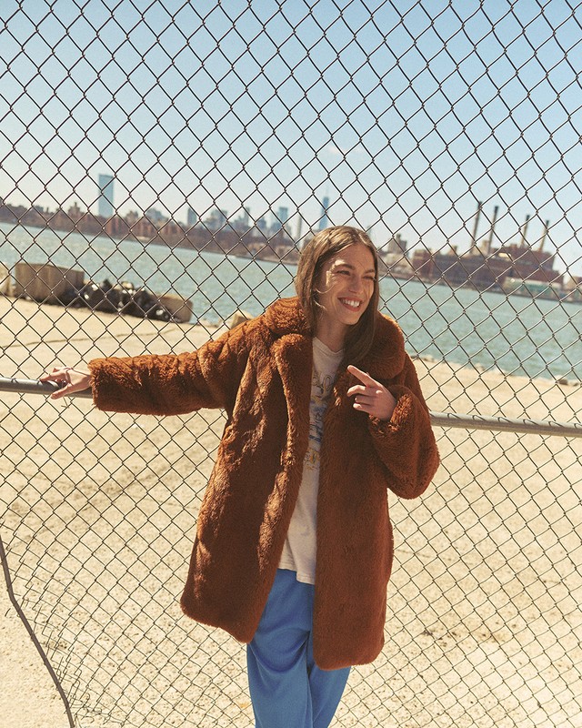 Woman in a brown faux fur coat poses in front of a chain-link fence with an urban waterfront and city skyline in the background.