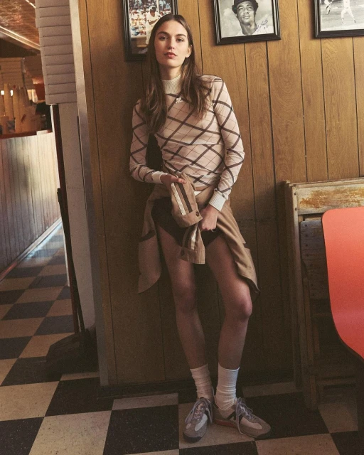 Young woman poses indoors by wood-paneled wall in patterned sweater, neutral skirt, tube socks, and sneakers, with framed photos on wall.