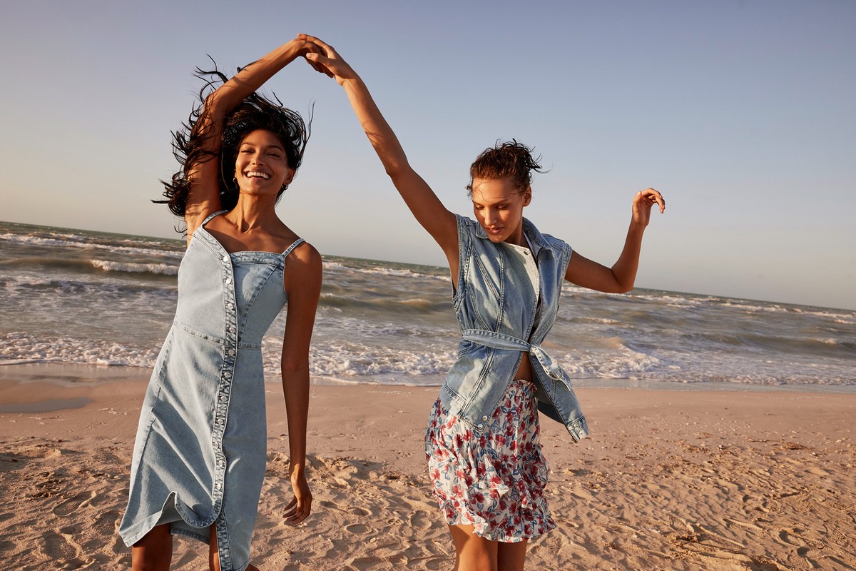 Two women joyfully dance on a sandy beach near the ocean, with one in a denim dress and the other in a floral skirt and denim vest.