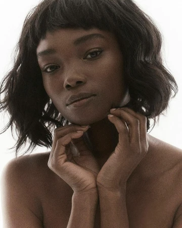 Close-up portrait of a Black woman with short dark wavy hair holding her hands under her chin against a bright background.