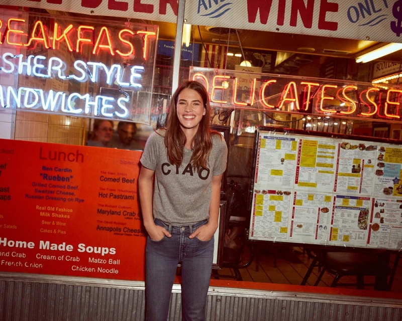 Woman in a gray t-shirt and blue jeans stands in front of a delicatessen with neon signs advertising breakfast, sandwiches, and wine.