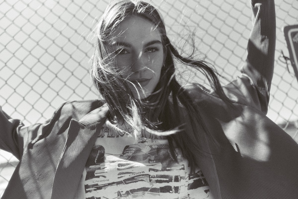 Black and white portrait of a young woman in a t-shirt and blazer, posed in front of a chain-link fence on a sunny day outdoors.