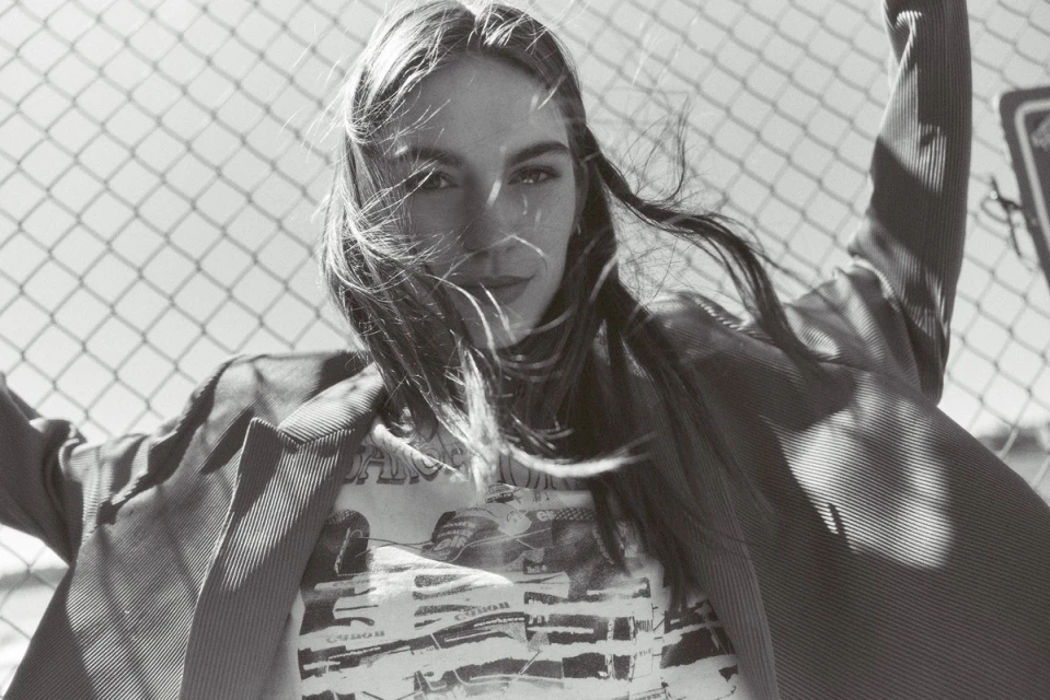 Black and white portrait of a young woman in a t-shirt and blazer, posed in front of a chain-link fence on a sunny day outdoors.