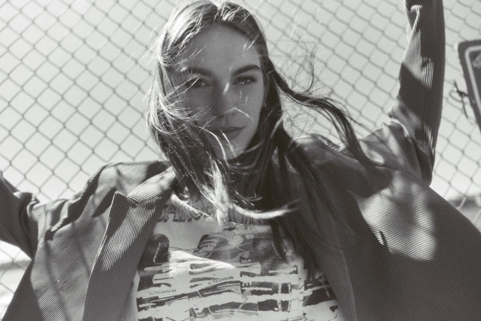 Black and white portrait of a young woman in a t-shirt and blazer, posed in front of a chain-link fence on a sunny day outdoors.