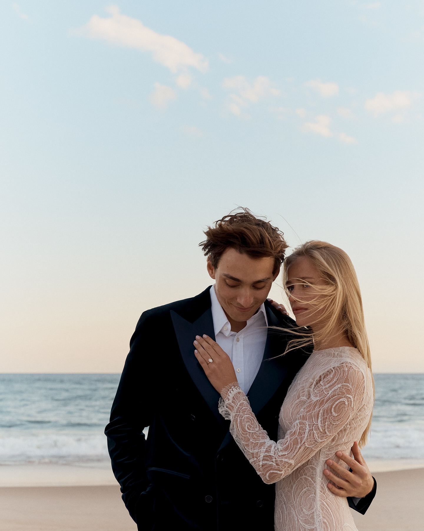 Romantic beach wedding photo: Couple in formal wear embraces by the ocean with a blue sky backdrop. Focus on love and celebration.