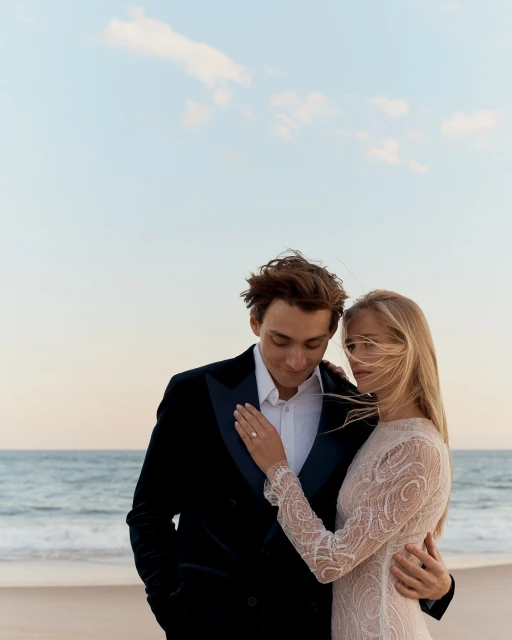Romantic beach wedding photo: Couple in formal wear embraces by the ocean with a blue sky backdrop. Focus on love and celebration.