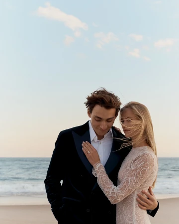 Romantic beach wedding photo: Couple in formal wear embraces by the ocean with a blue sky backdrop. Focus on love and celebration.