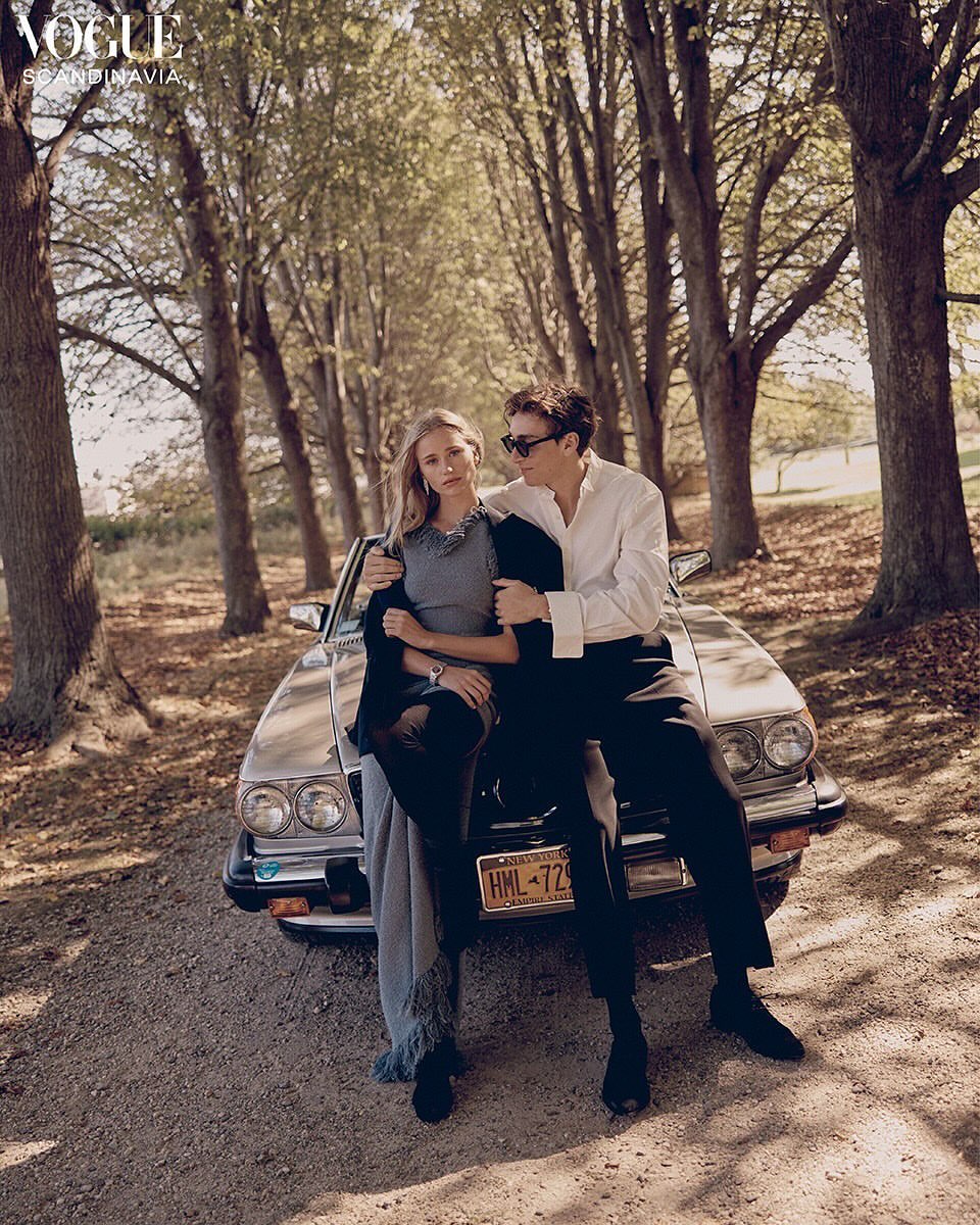 Man and woman sitting on the hood of a vintage car parked on a tree-lined dirt road, photographed by Landon Mcmahon.