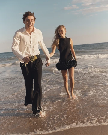 A happy couple is walking barefoot through the waves on a sunny beach, wearing elegant outfits and holding hands, enjoying the seaside.