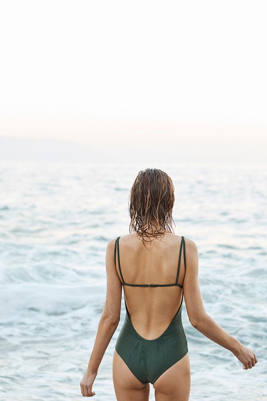 A woman in a swimsuit stands facing the ocean with her back to the camera in a sunlit, outdoor beach setting.