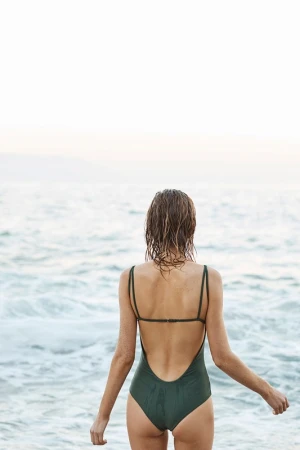 A woman in a swimsuit stands facing the ocean with her back to the camera in a sunlit, outdoor beach setting.