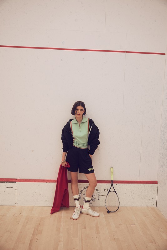 A young woman poses in a squash court wearing a matching mint green top and black shorts, holding a red towel next to a racket.