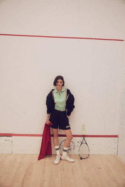 A young woman poses in a squash court wearing a matching mint green top and black shorts, holding a red towel next to a racket.