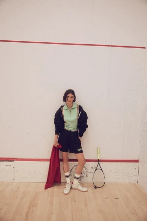 A young woman poses in a squash court wearing a matching mint green top and black shorts, holding a red towel next to a racket.