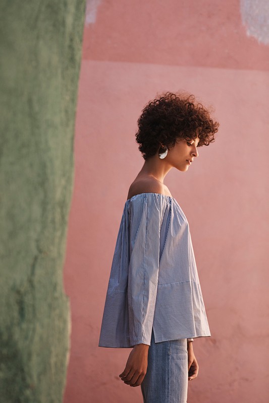 Profile portrait of a woman with curly hair wearing an off-the-shoulder striped top against a pink wall