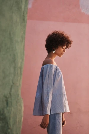 Profile portrait of a woman with curly hair wearing an off-the-shoulder striped top against a pink wall