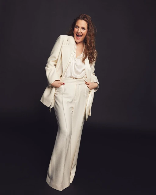 Drew Barrymore in a white suit posing against a dark gray background with an open mouth and a cheerful expression, full shot.