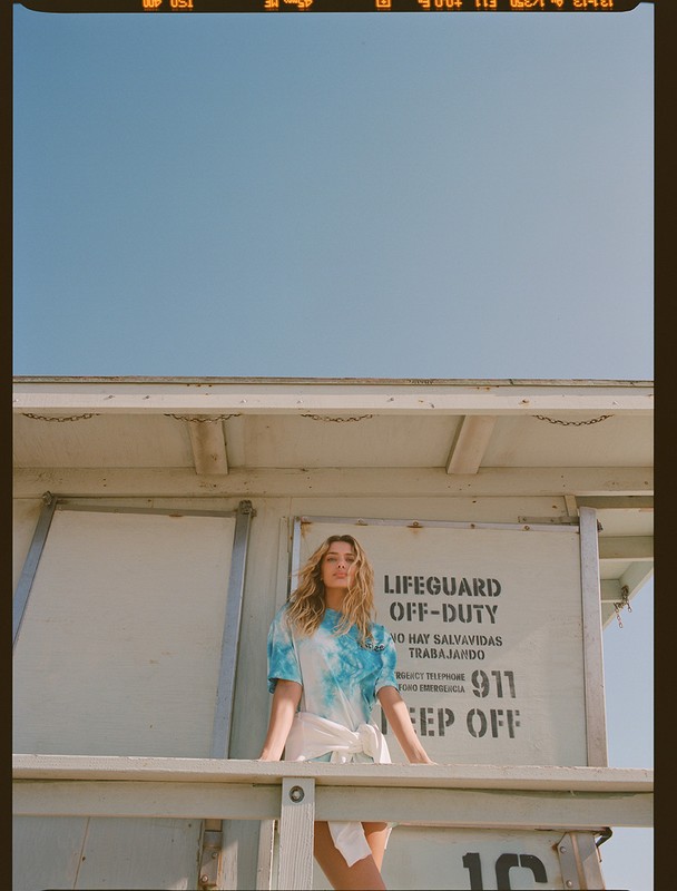 A woman stands on a lifeguard station against a clear sky, wearing a tie-dye shirt and white skirt, captured with natural light.