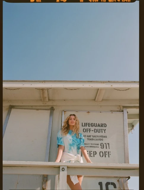 A woman stands on a lifeguard station against a clear sky, wearing a tie-dye shirt and white skirt, captured with natural light.